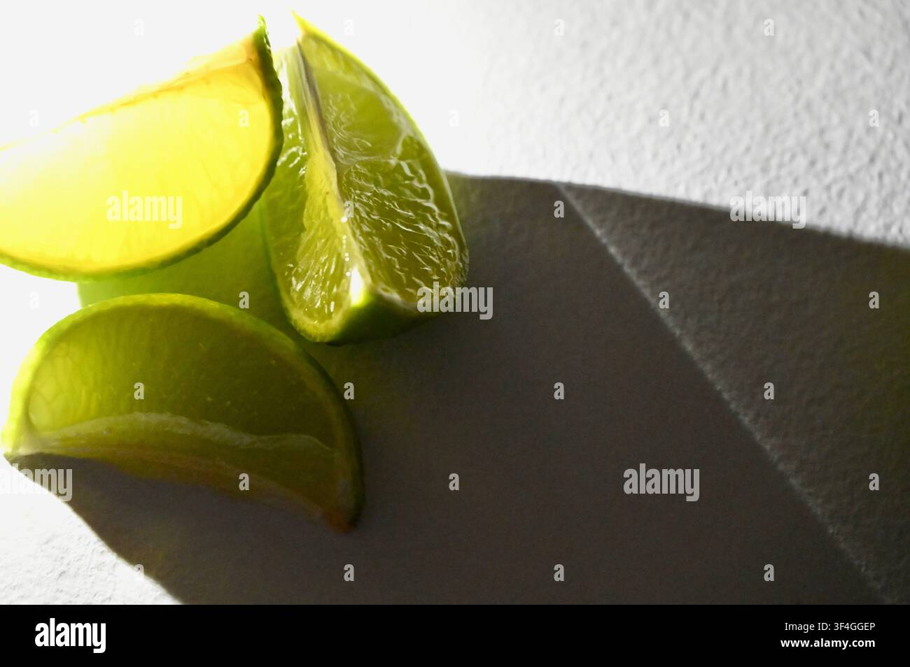 Una fotografia macro ad alto contrasto con fresche scorze di lime verdi su uno sfondo bianco texture. La luce solare diritta e luminosa illumina la polpa succosa degli agrumi, creando ombre geometriche nette e scure sulla superficie. Una composizione moderna e minimalista perfetta per la fotografia alimentare, ricette di bevande estive, concetti di cocktail, menu del bar e branding degli agrumi freschi. Foto Stock