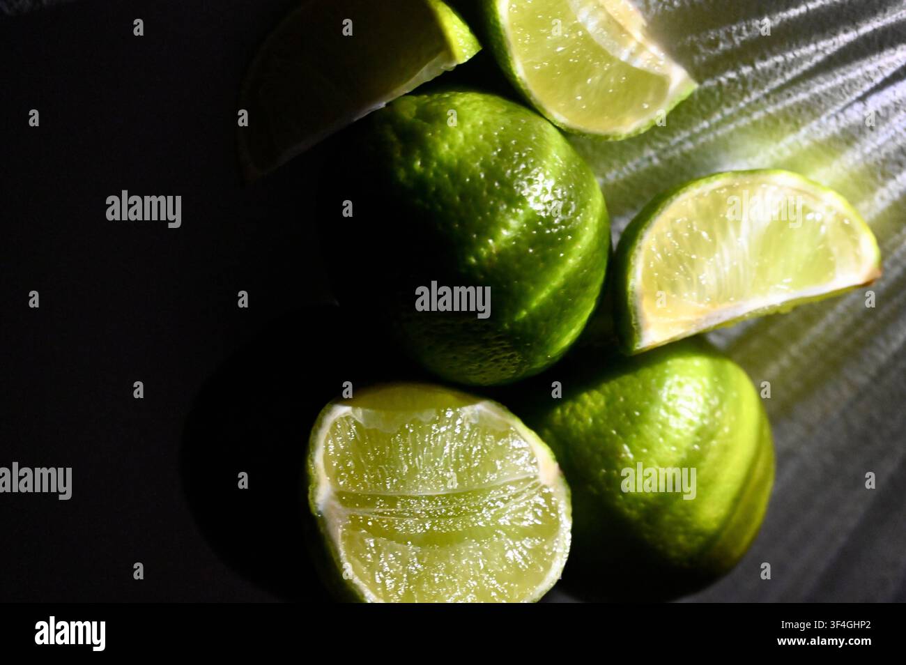 Una fotografia macro cupa e a bassa chiave con lime verdi interi insieme a metà e spicchi freschi su uno sfondo scuro. Una forte illuminazione direzionale crea riflessi luminosi sulla buccia strutturata e sulla polpa succosa, mentre proietta ombre profonde e drammatiche. Immagine perfetta per menu di bar cocktail, branding degli agrumi, fotografia alimentare, concetti di bevande tropicali e design culinario. Foto Stock