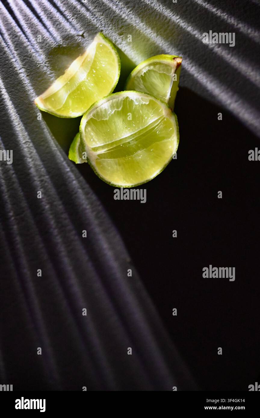 Una fotografia macro cupa e a bassa chiave con lime verdi interi insieme a metà e spicchi freschi su uno sfondo scuro. Una forte illuminazione direzionale crea riflessi luminosi sulla buccia strutturata e sulla polpa succosa, mentre proietta ombre profonde e drammatiche. Immagine perfetta per menu di bar cocktail, branding degli agrumi, fotografia alimentare, concetti di bevande tropicali e design culinario. Foto Stock