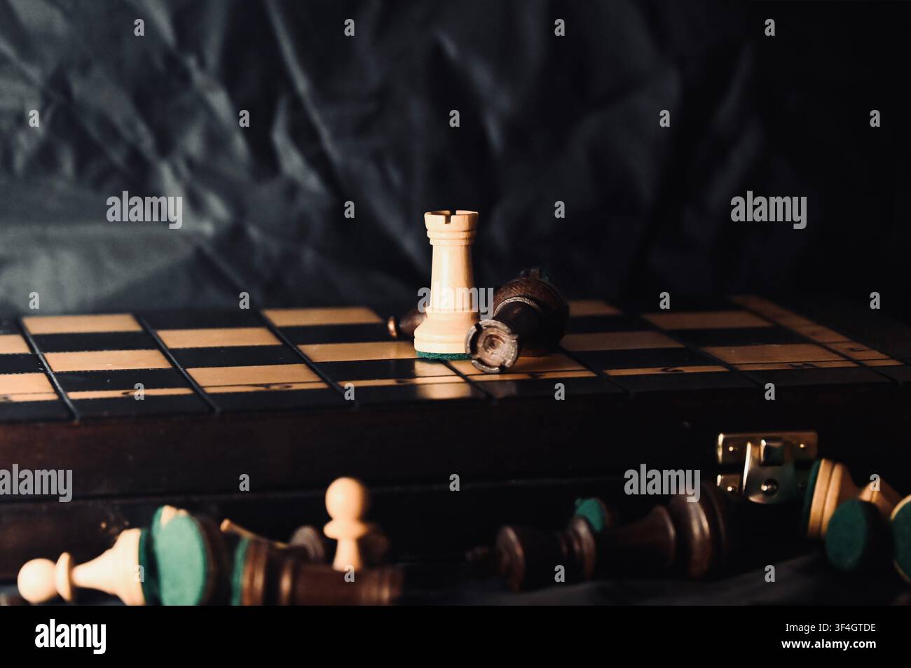 Una fotografia cupa e sobria di una classica configurazione di scacchi in legno su uno sfondo scuro. Un singolo alfiere bianco rimane in piedi sull'ascrizione, circondato da pezzi di scacchi scuri e chiari caduti, simboleggiando vittoria strategica, sopravvivenza, resilienza e competizione commerciale. Foto Stock