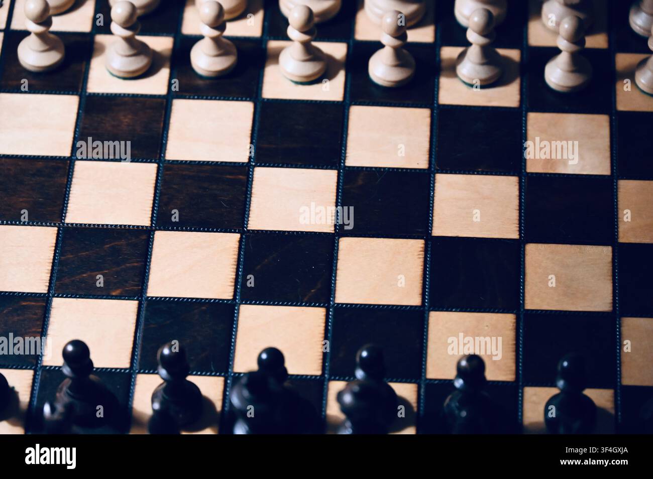 Vista ad angolazione alta di una scacchiera di legno con tutte le pedine disposte in posizione di partenza. Il contrasto drammatico tra tonalità chiare e scure con ombre sottili simboleggia la pianificazione strategica, la competizione aziendale, la visione tattica, il processo decisionale e la sfida intellettuale. Foto Stock