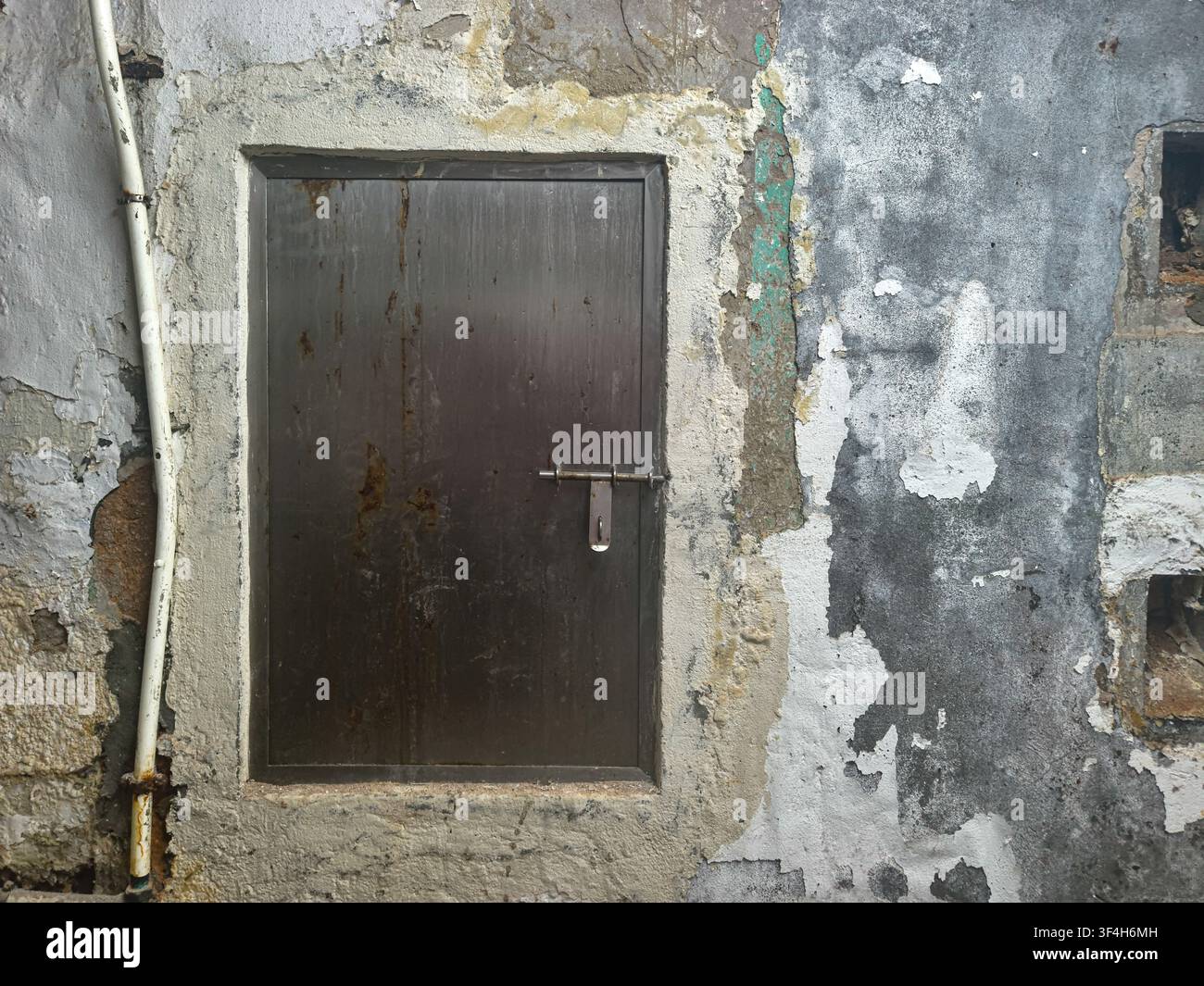 piccola porta di servizio in metallo logorata, fissata con una serratura a slitta tradizionale e un lucchetto appeso su un muro di muratura fatiscente con vernice sbiadita Foto Stock
