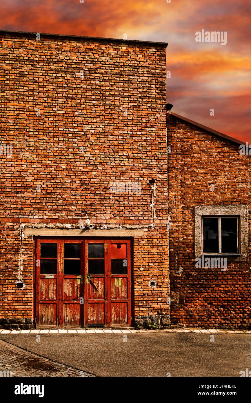 Edificio industriale in mattoni rossi stagionato con porte e finestre consumate sotto un drammatico cielo arancione al tramonto Foto Stock