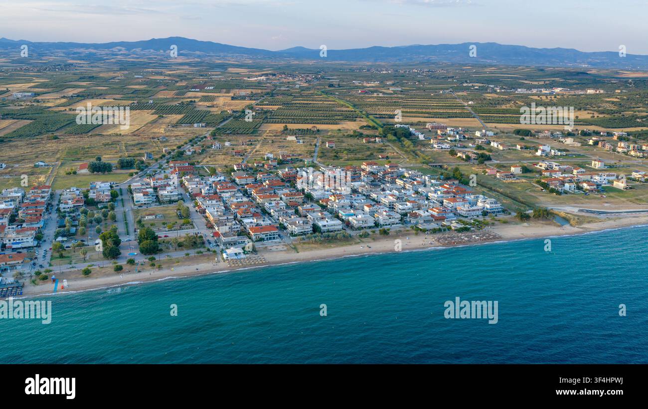 Una vista elevata mostra le case costiere compatte di Paralia Dionisiou, la riva sabbiosa, i campi coltivati e le basse montagne sulla costa greca. Foto Stock