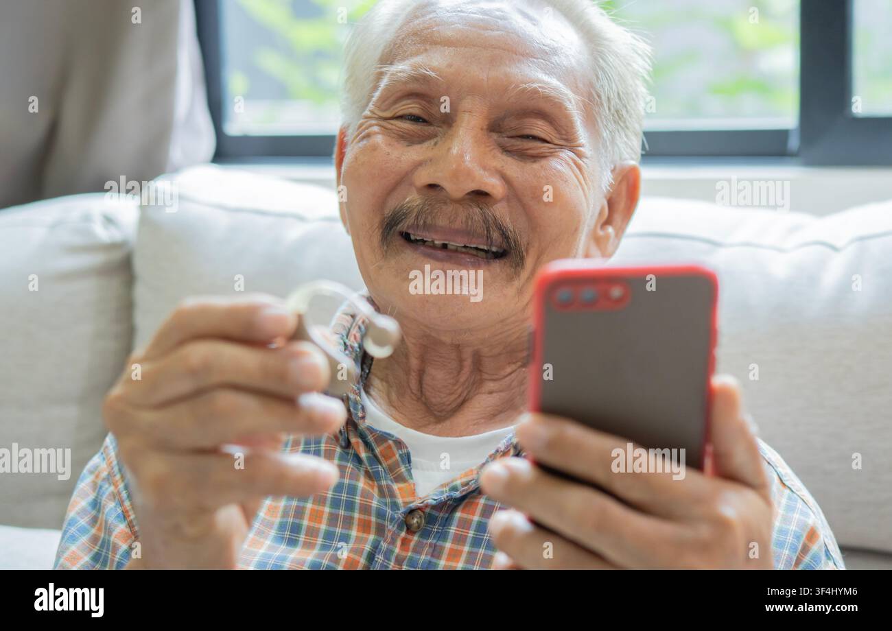 Uomo anziano felice che sorride mentre mostra un apparecchio acustico e un telefono, sottolineando l'importanza della salute uditiva e della tecnologia assistenziale innovativa per Foto Stock