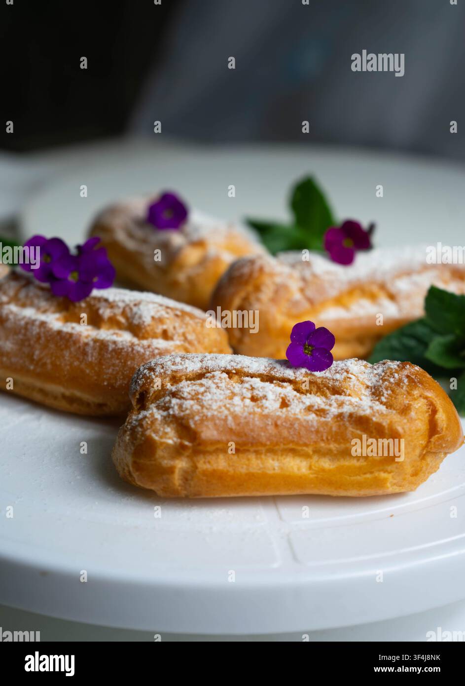 Éclair ripieni di crema fresca spolverati di zucchero a velo e decorati con fiori commestibili. Pasticceria francese tradizionale servita su un piatto bianco. Foto Stock