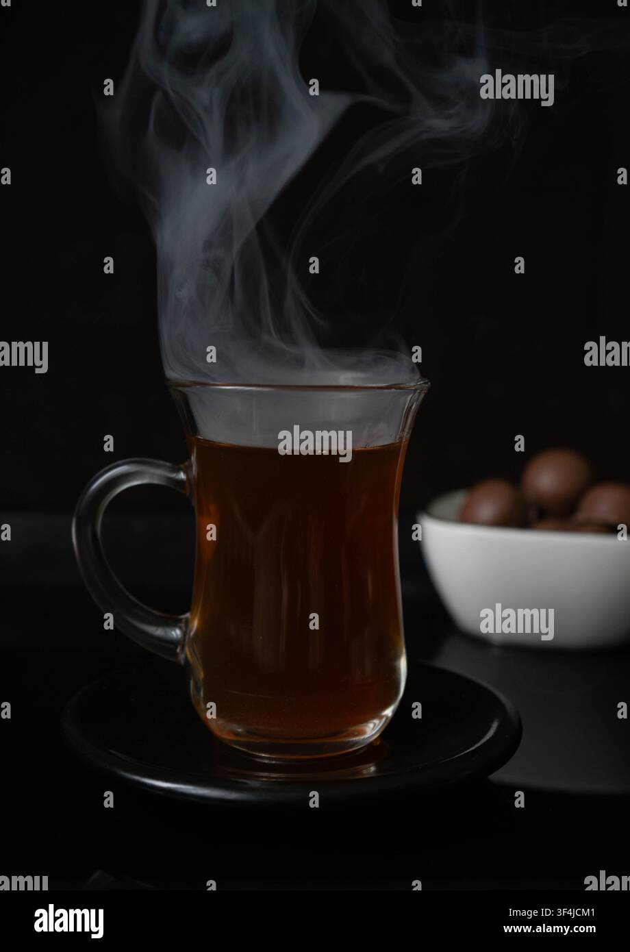 Tè nero caldo servito in una tazza di vetro tradizionale con vapore drammatico su uno sfondo scuro. Concetto di bevanda calda e accogliente. Foto Stock