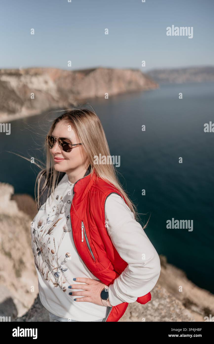 Donna viaggio costiero donna bionda in giacca rossa in piedi su una scogliera costiera che si affaccia sull'oceano blu durante una giornata luminosa Foto Stock