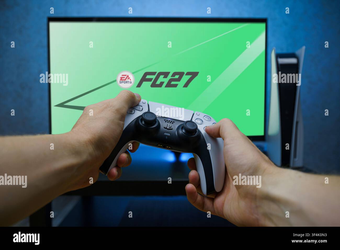 Mani che tengono un controller PlayStation 5 DualSense davanti a uno schermo TV che mostra il gioco EA Sports FC 27, concetto di gaming su console e videogioco di calcio. Foto Stock