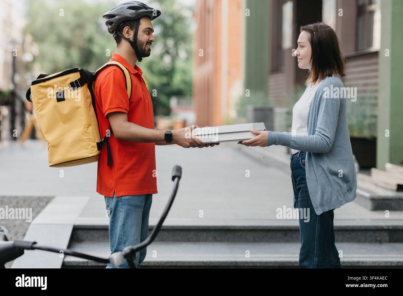 Ordine pranzo. La ragazza prende scatole di pizza dal corriere in casco con zaino giallo e bicicletta Foto Stock
