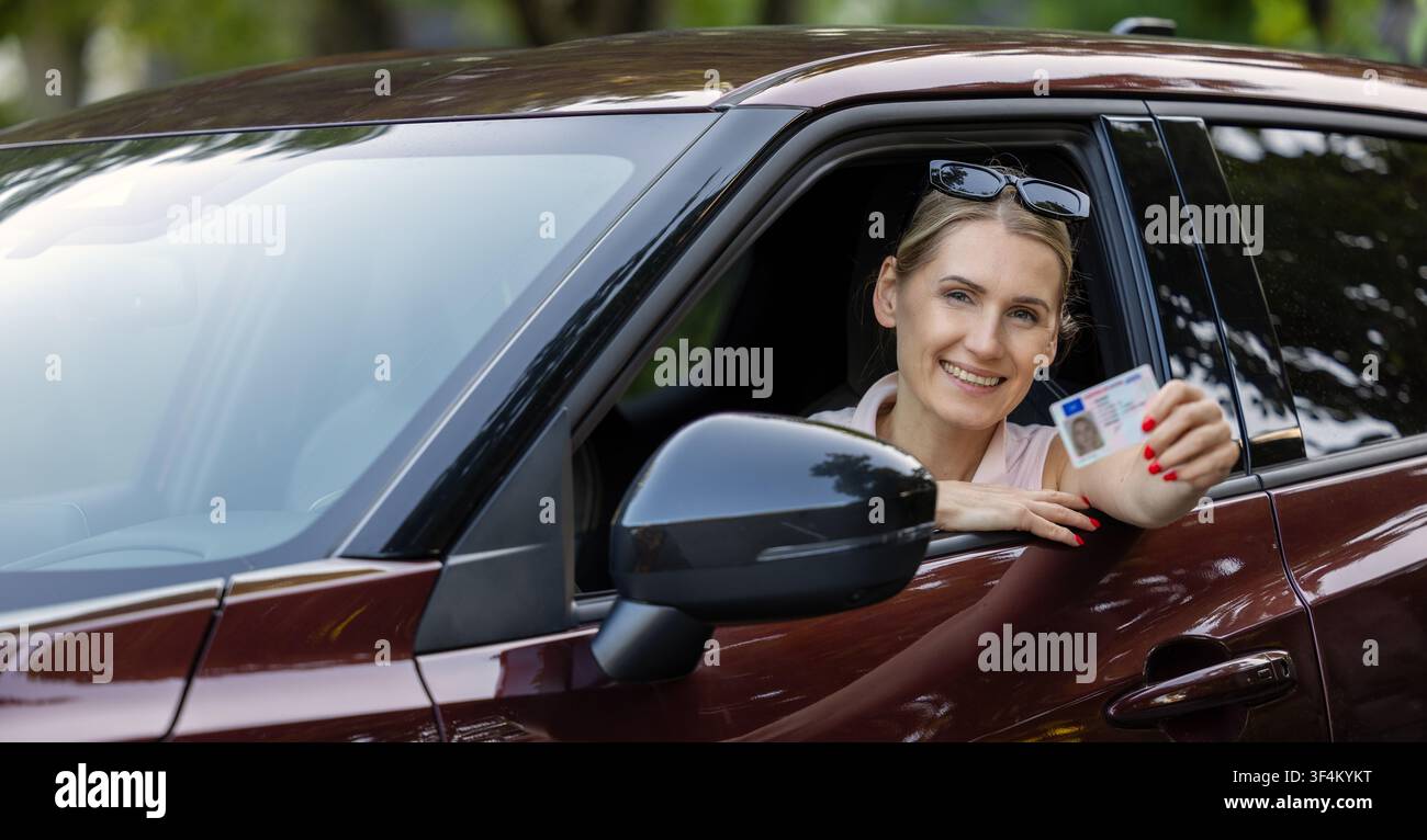 Donna sorridente seduta in auto e che mostra il suo nuovo patentino di guida fuori dal finestrino alla telecamera Foto Stock