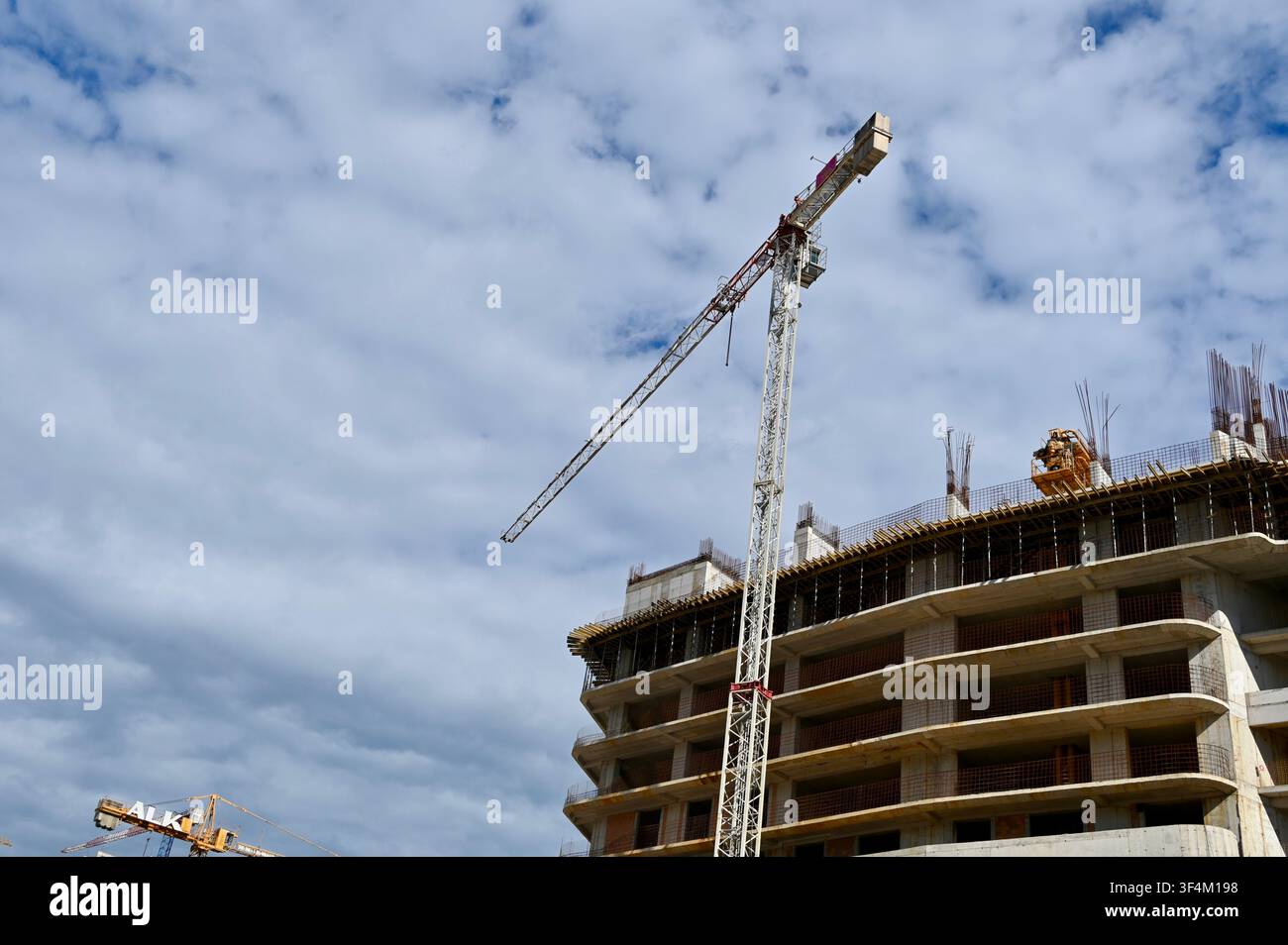 Una ripresa di una grande gru torre di costruzione che opera accanto a un moderno edificio residenziale a più piani in costruzione sotto un cielo blu nuvoloso. Ideale per concetti di sviluppo immobiliare, architettura, ingegneria e crescita urbana. Foto Stock