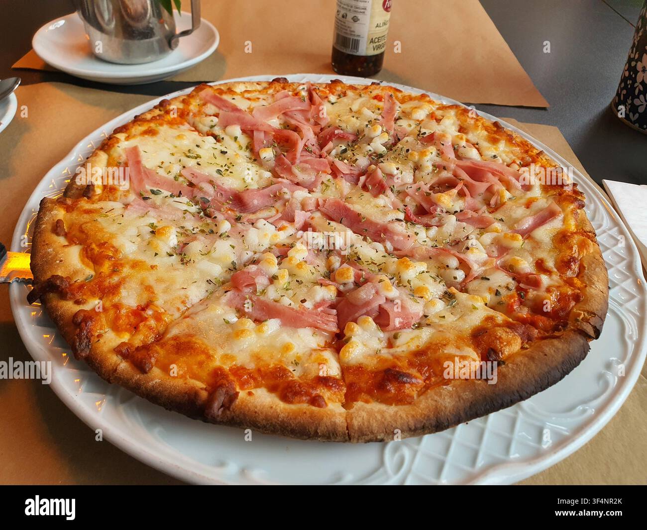 Una pizza intera rotonda servita su un piatto decorativo in ceramica bianca, ricoperta di mozzarella fusa, fette di prosciutto rosa, chicchi di mais dolce e Foto Stock