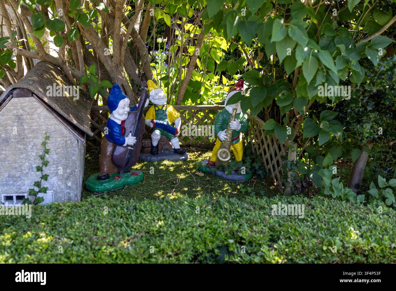 Statue colorate di gnomi da giardino che suonano strumenti musicali accanto a una casetta in miniatura sotto alberi frondosi in un giardino paesaggistico che crea un'atmosfera capricciosa Foto Stock