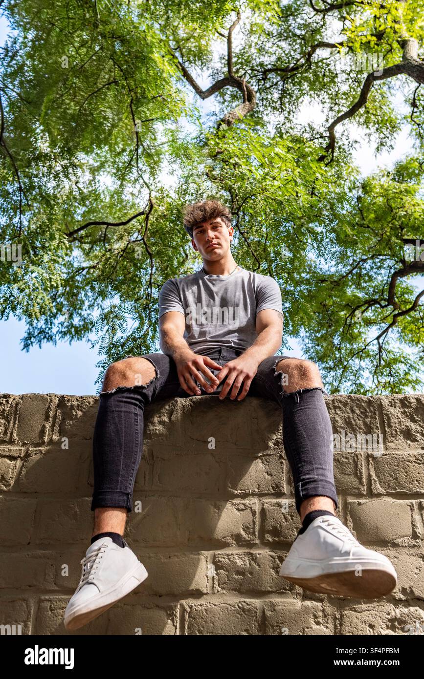 Ritratto di stile urbano di un uomo Gen Z seduto in alto su un muro che guarda in basso verso la fotocamera Foto Stock