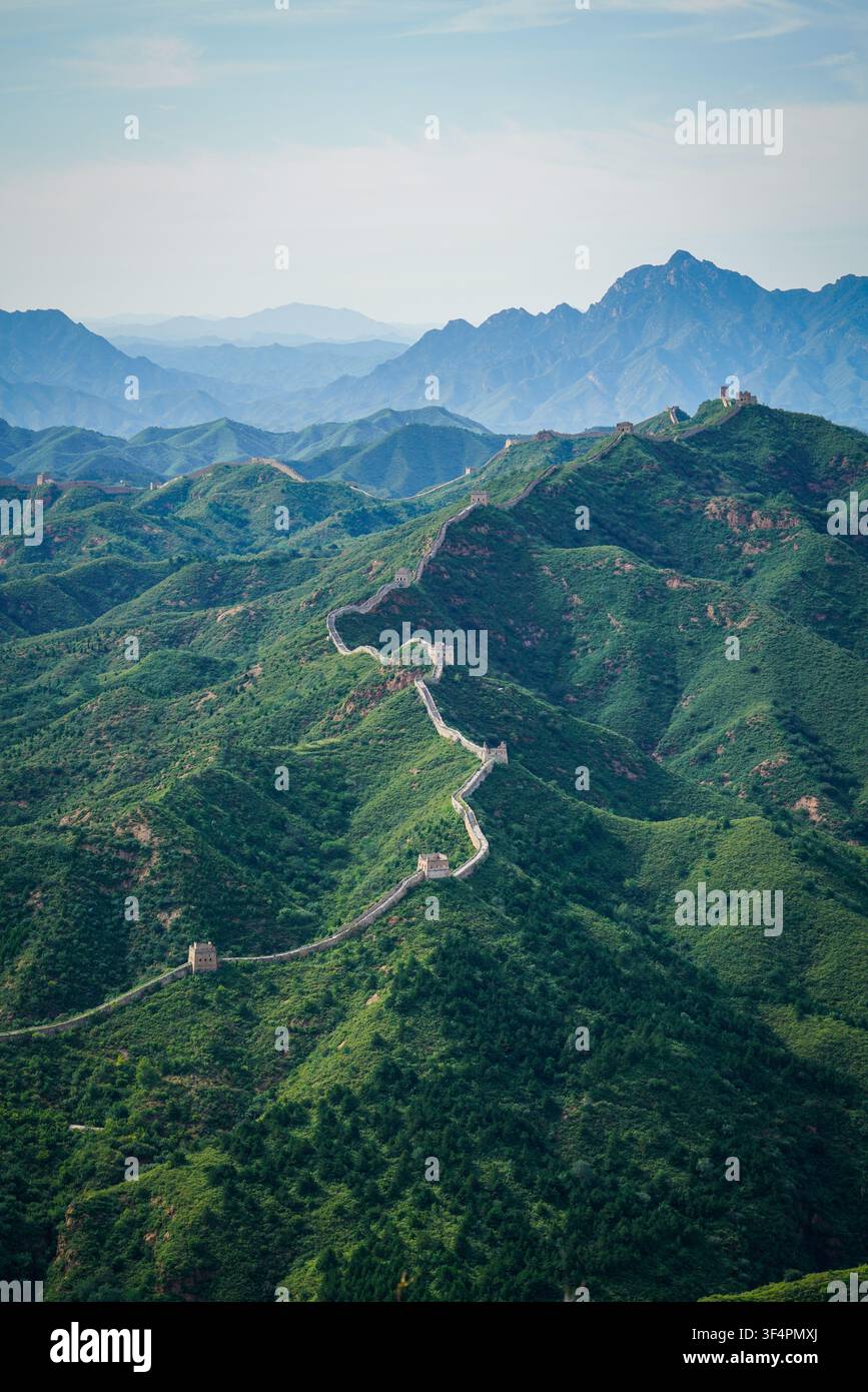 Un segmento della Grande Muraglia della Cina in pietra grigia con diverse torri di guardia si snoda attraverso una serie di montagne ripide, verdi e ricoperte di alberi sotto un b pallido Foto Stock