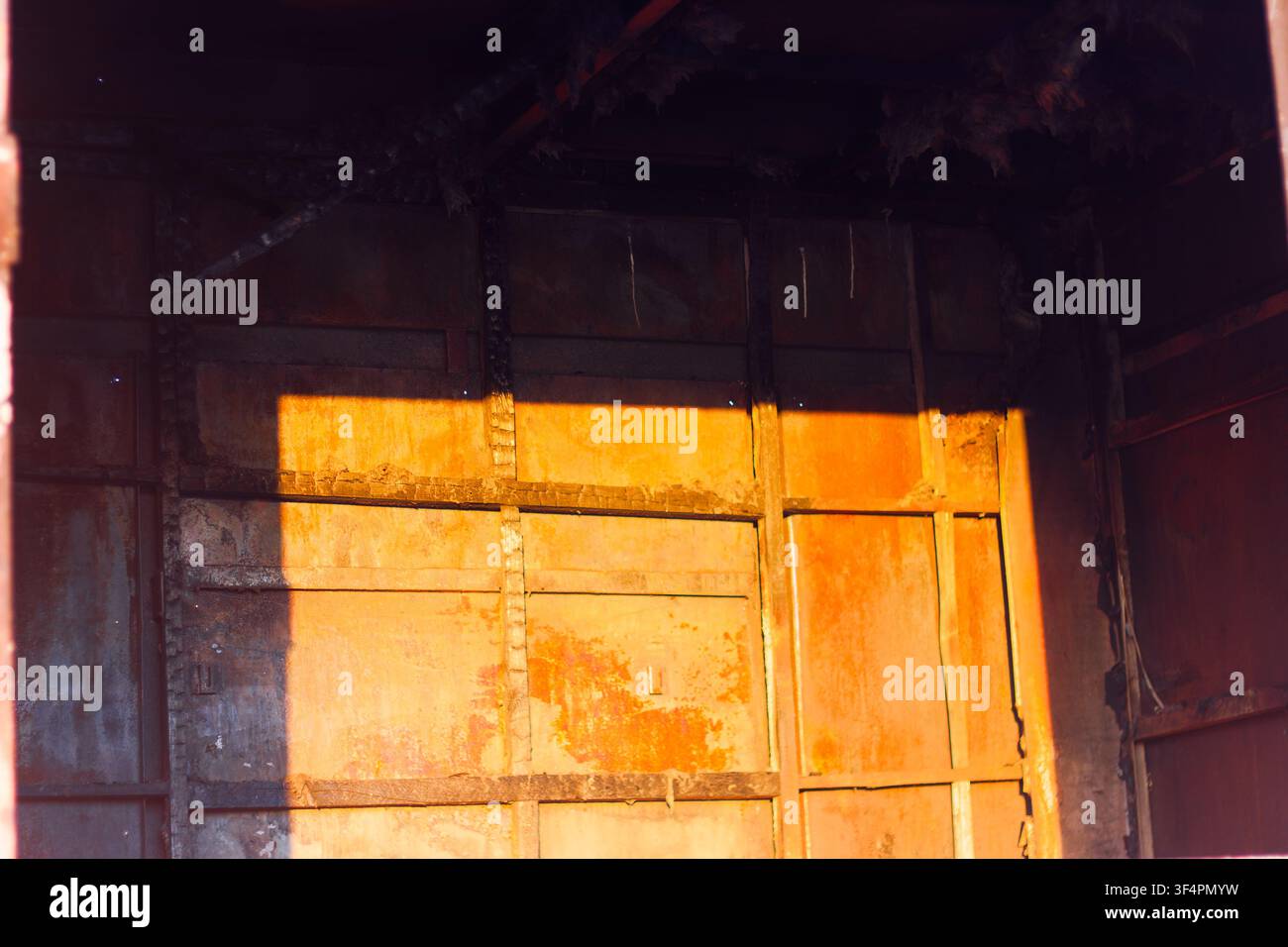 Interno in metallo bruciato di un capannone abbandonato illuminato dalla luce dorata del sole, che mostra muri carbonizzati e resti strutturali di una cabina utilitaria distrutta a Foto Stock