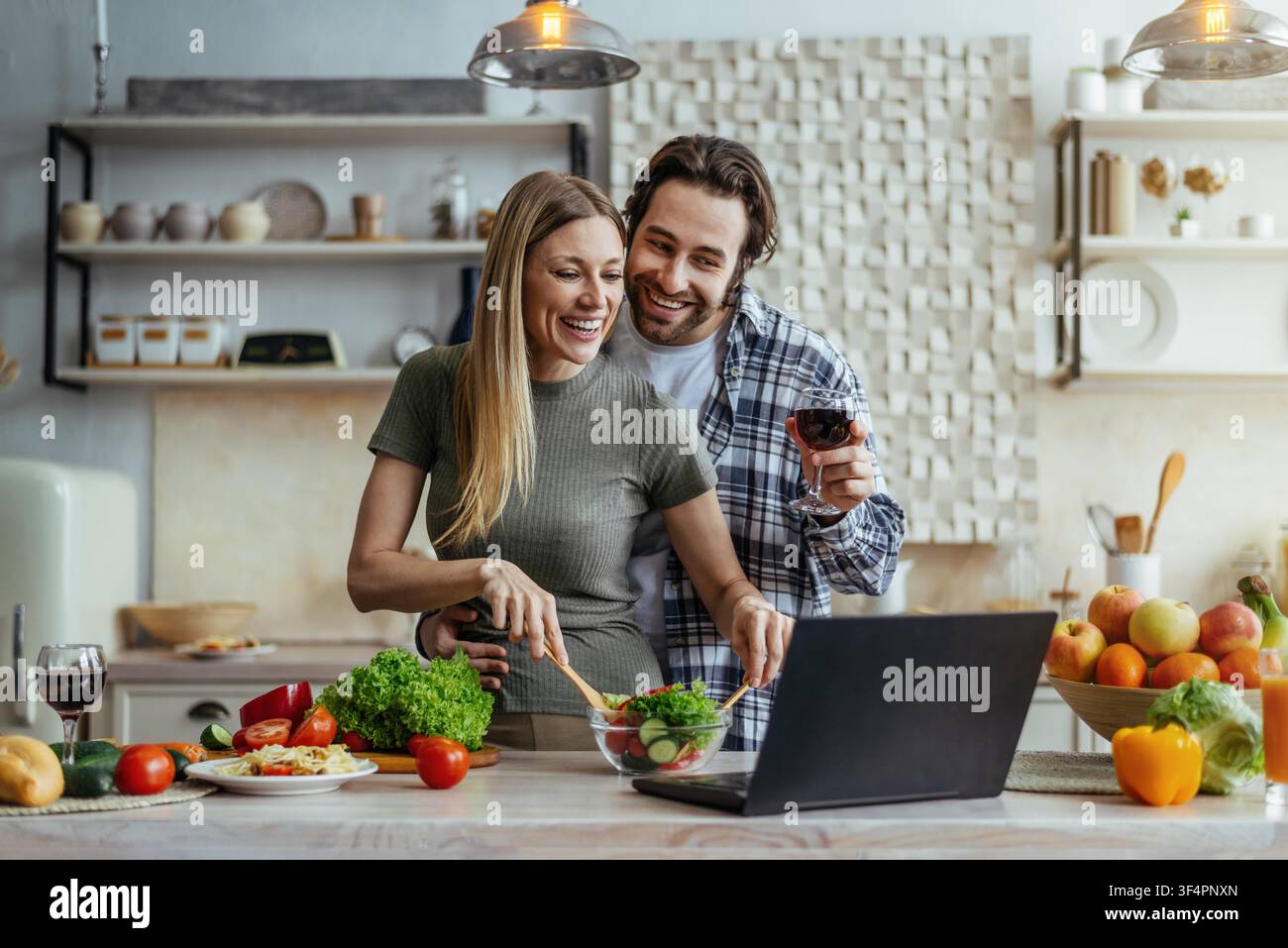 Giovane coppia felice che cucina insieme in cucina, prepara un'insalata sana con laptop, concetto organico Foto Stock