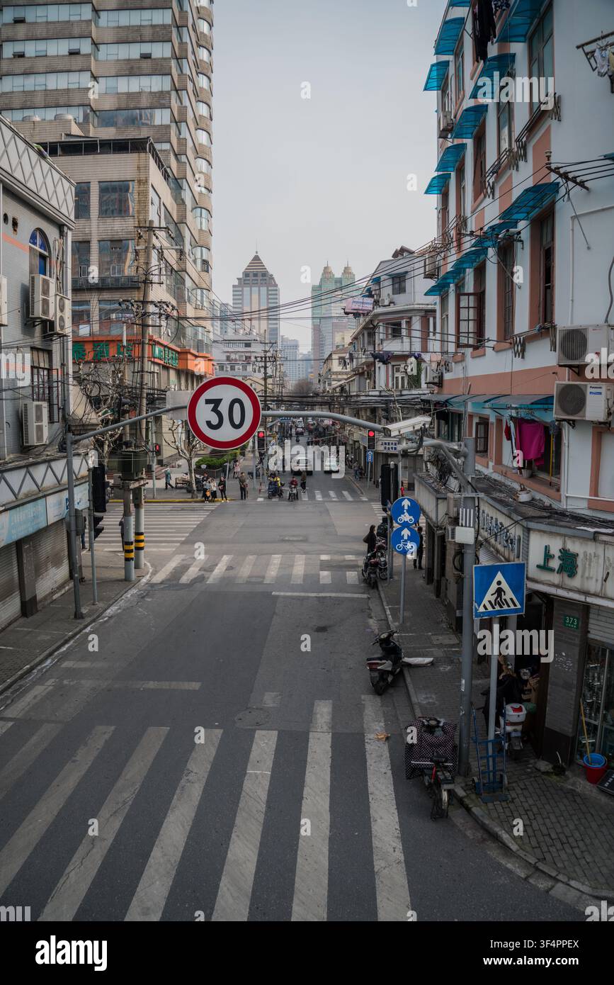 Una stretta strada lastricata di grigio con attraversamenti pedonali bianchi si estende nello sfondo, fiancheggiata da edifici di altezze ed età diverse sotto un ove Foto Stock