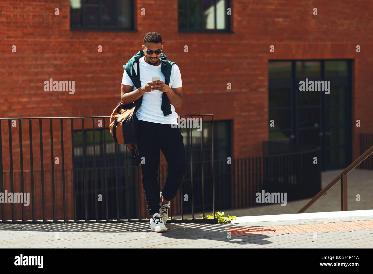 Giovane uomo nero che scrive al telefono mentre si appoggia a una ringhiera, in un ambiente all'aperto Foto Stock