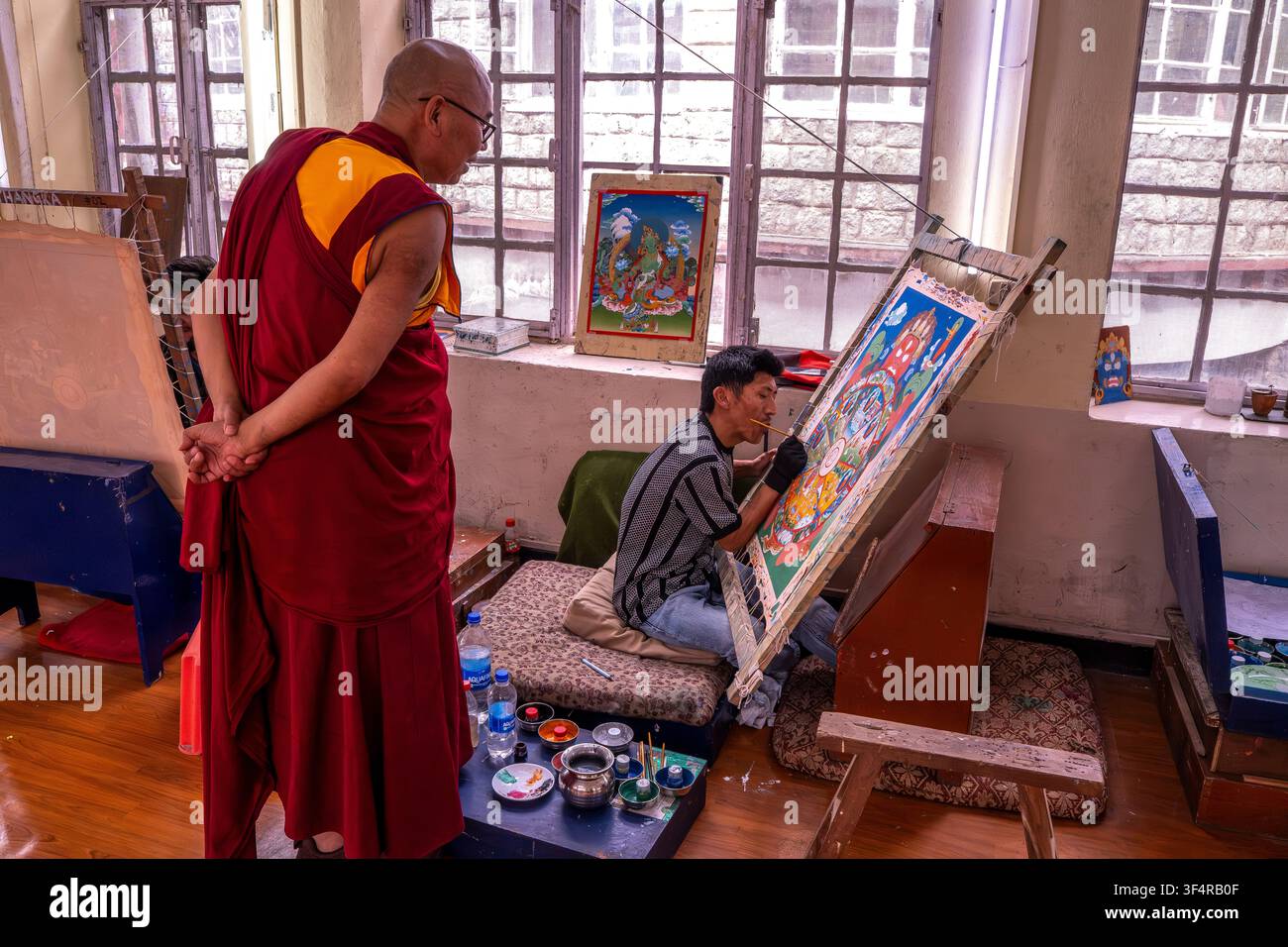 Istituto Norbulingka, Sidhpur, Himachal Pradesh, India. Un monaco osserva un artista dipingere un thangka in uno studio. L'artista è seduto e concentrato sul suo Foto Stock