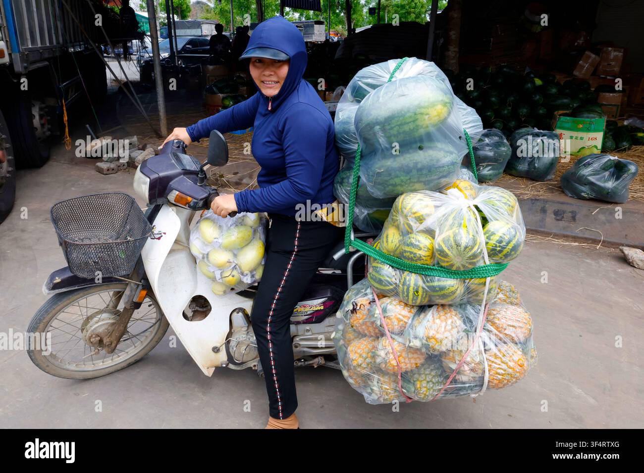 Una donna sta trasportando una grande quantità di frutta e verdura sulla sua moto. Il carico è impressionante, composto da angurie, meloni cantalupo, un Foto Stock
