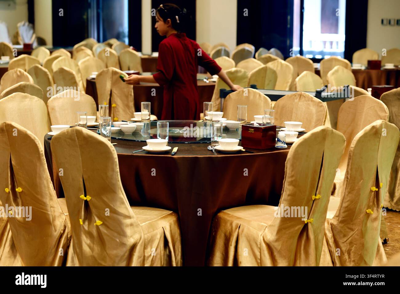 Una cameriera allestisce una sala da banchetto con tavoli preparati per un evento. La sala è decorata con sedie drappeggiate e posate. Hoa Lu, Vietnam. Foto Stock