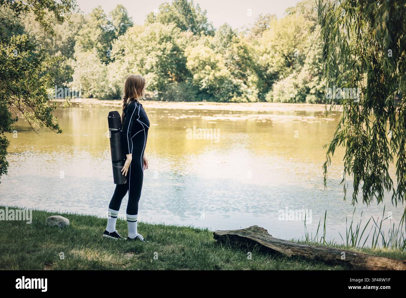Donna con tappetino da yoga in piedi vicino al lago, passeggiata contemplativa nella natura, viaggio di fitness in solitaria, motivazione per allenamenti scenografici, pace e solitudine Foto Stock