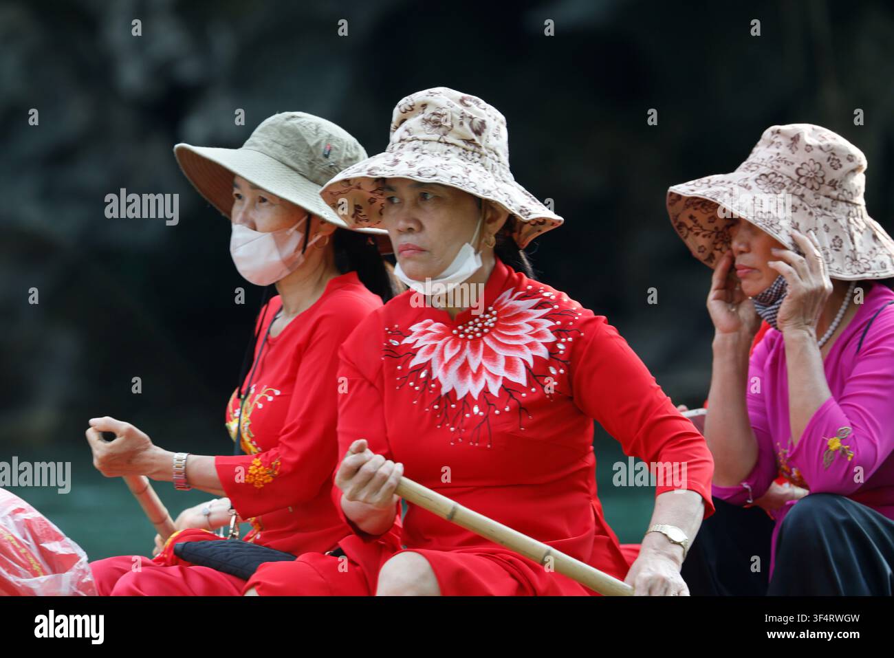 Tre donne con cappelli e mascherine navigano su una barca. Sono vestite con abiti tradizionali rossi e rosa e si godono una giornata di sole. Trang An Foto Stock