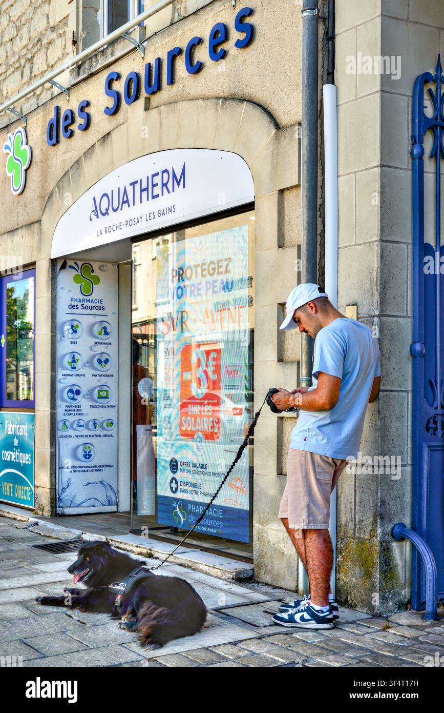 Uomo vestito in modo casual appoggiato al muro, naviga sul cellulare mentre tiene un cane a riposo al guinzaglio – La Roche Posay, Vienne (86), Francia. Foto Stock