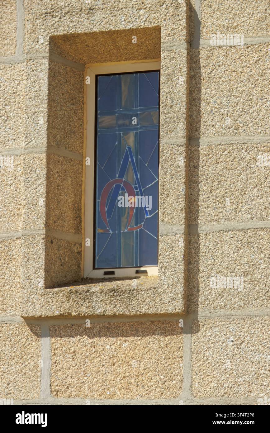 Una finestra stretta in un muro di pietra granito rustico con una vetrata blu che mostra simboli teologici cristiani Alpha e Omega Foto Stock