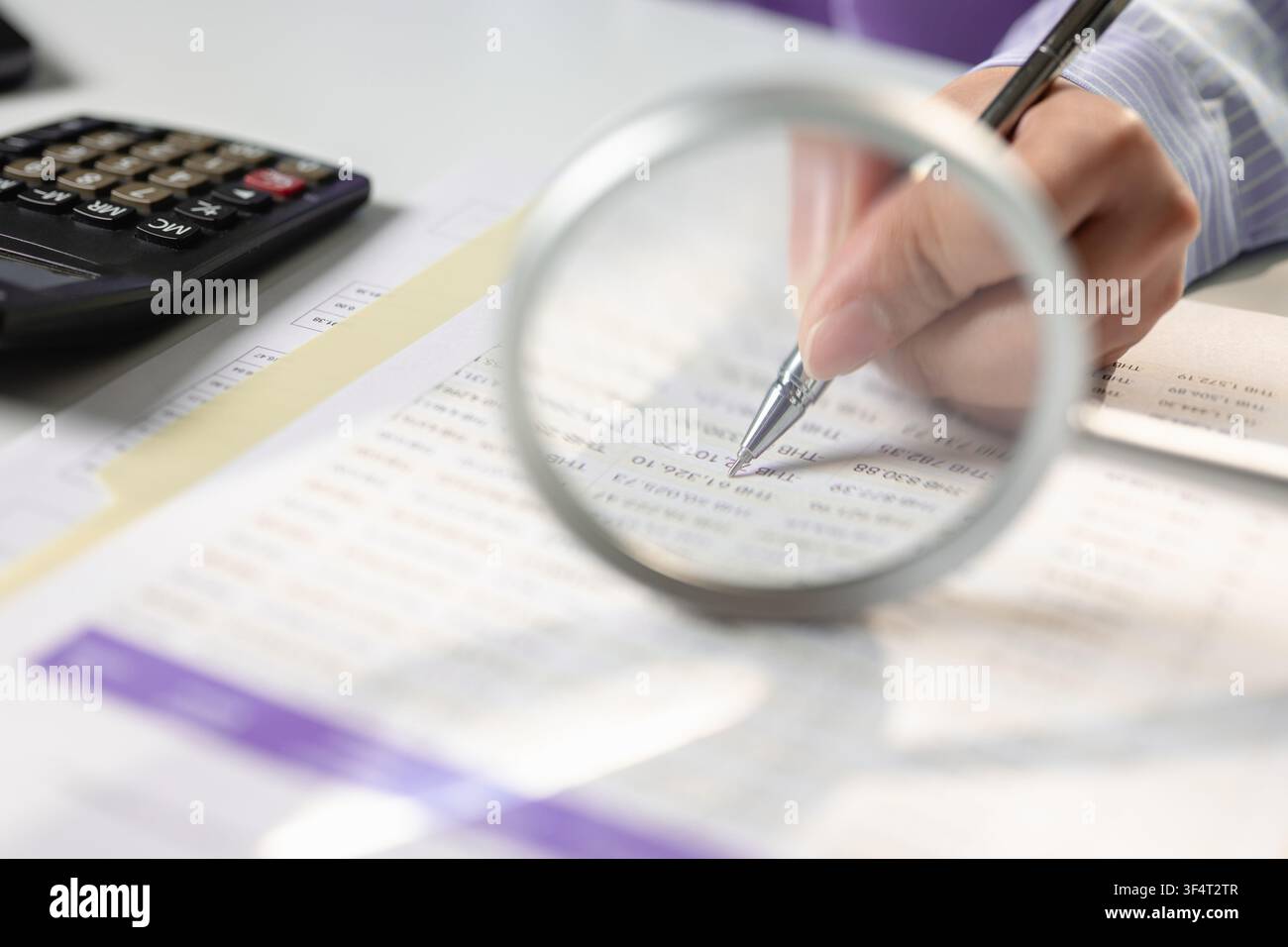Prima piano delle mani di un revisore che tiene una penna e ispeziona un documento di rapporto finanziario attraverso una lente d'ingrandimento sulla scrivania. Concetto di processo di revisione fiscale, budg Foto Stock