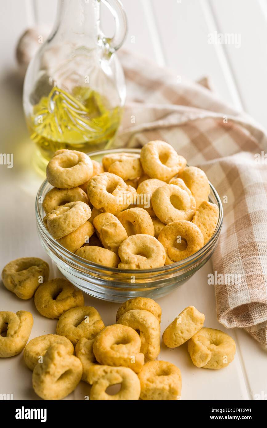 Cracker taralli dorati a forma di anello ammucchiati in una ciotola di vetro accanto a una bottiglia di olio d'oliva e un canovaccio piegato, che trasmettono una rustica e croccante Medit Foto Stock
