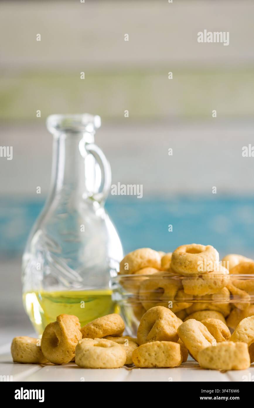 Cracker taralli dorati a forma di anello ammucchiati in una ciotola di vetro accanto a una bottiglia di olio d'oliva e un canovaccio piegato, che trasmettono una rustica e croccante Medit Foto Stock
