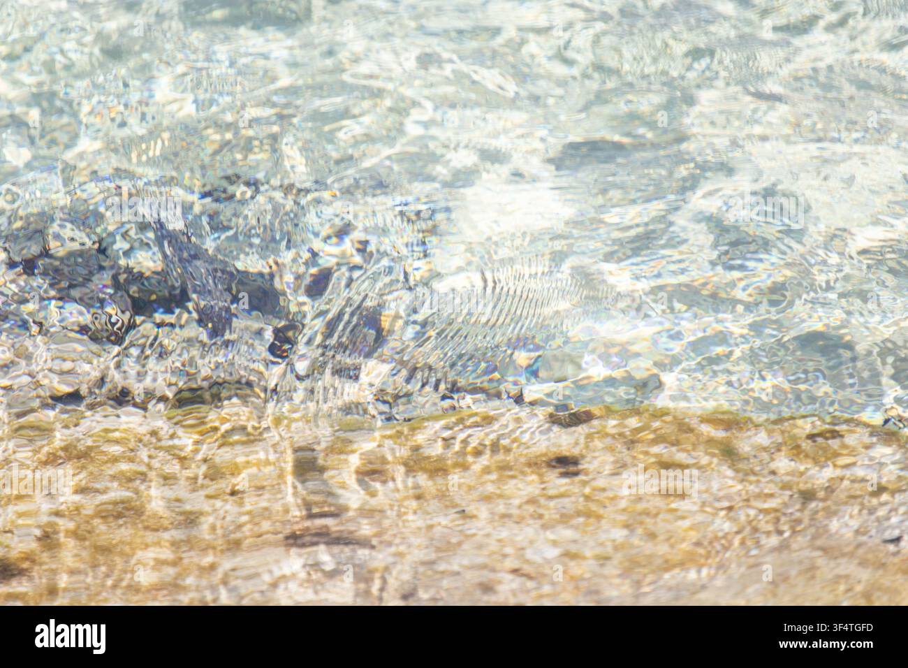 Acqua di mare cristallina che ondeggia su un fondale sabbioso e roccioso in una giornata di sole, texture astratta della superficie dell'acqua naturale e riflessi di luce Foto Stock