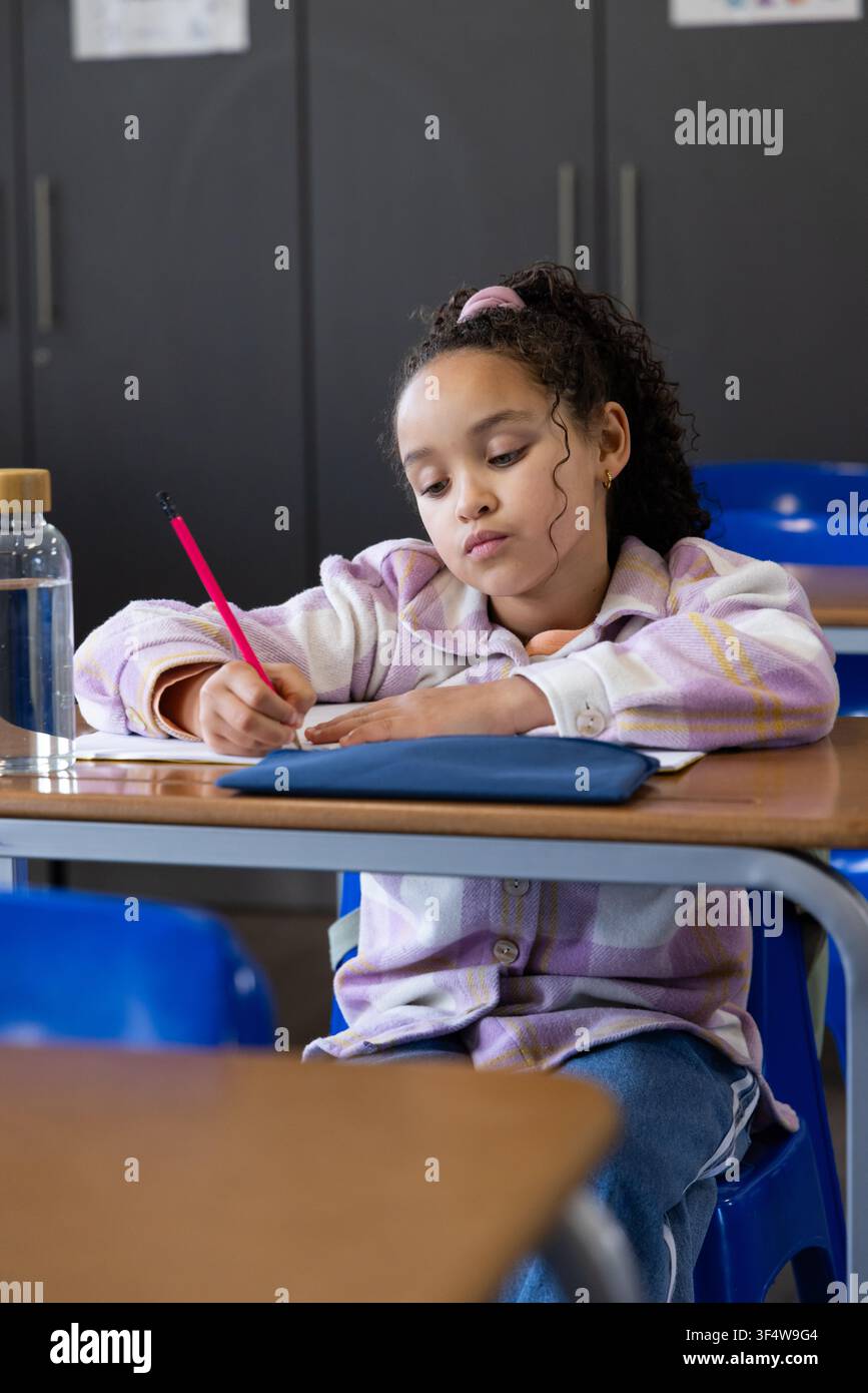 Scolaretta afroamericana che scrive in classe, tiene una matita rossa, un quaderno e un astuccio blu Foto Stock