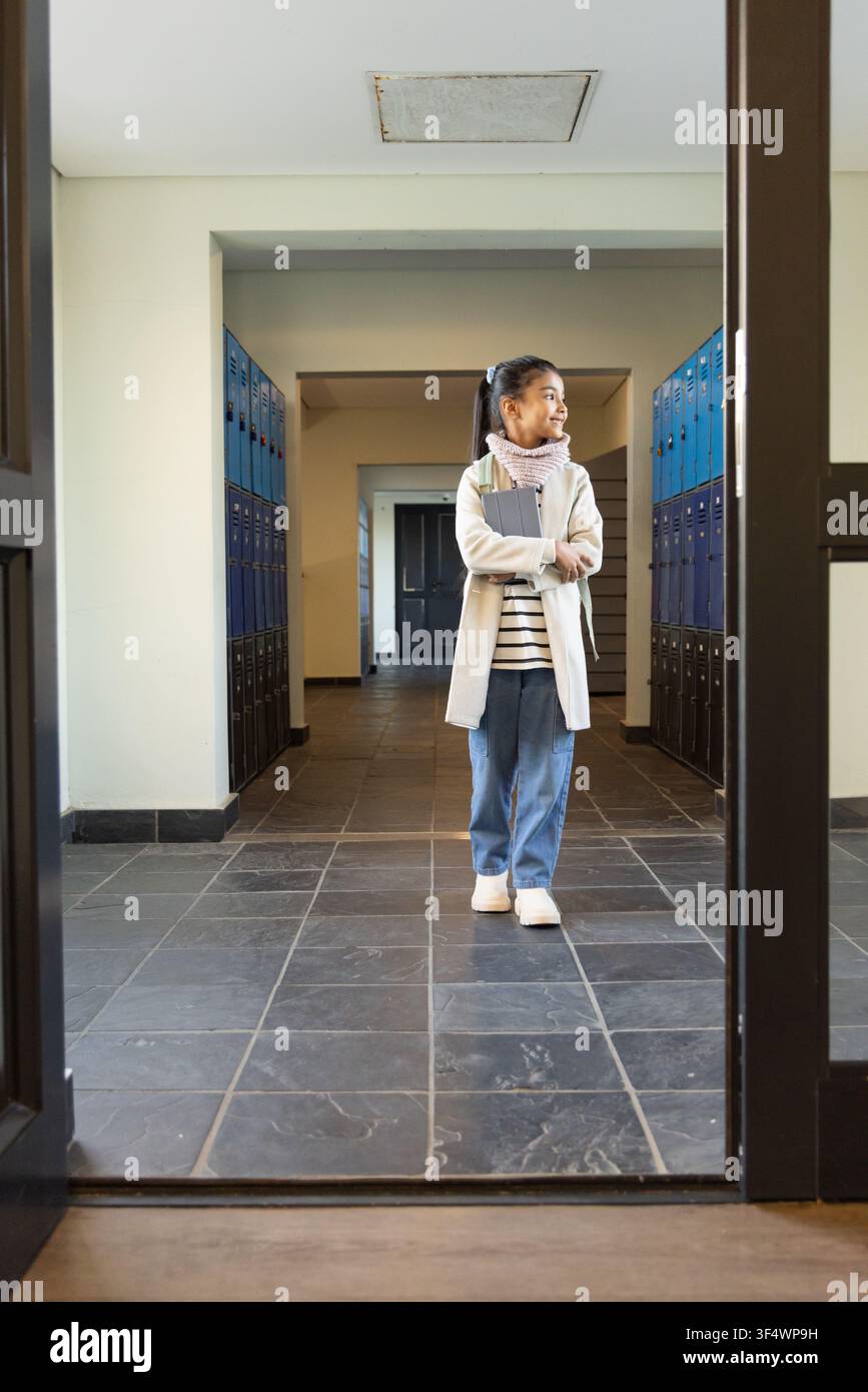 Studentessa adolescente con cappotto e zaino, in piedi nella sala della scuola con raccoglitore e armadietti Foto Stock