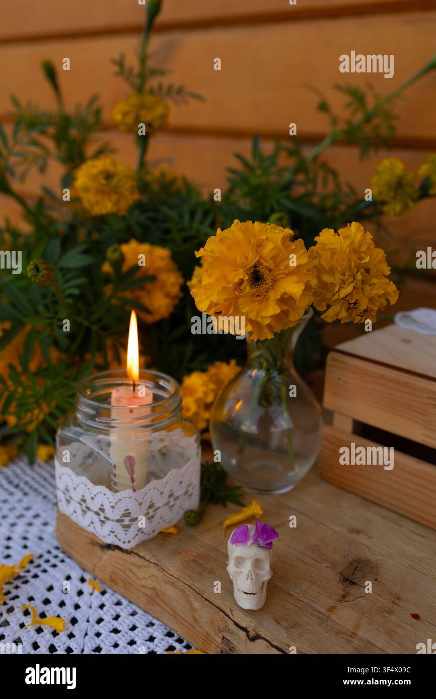 Primo piano di fiori di calendula arancioni brillanti in vasi di vetro accanto a una candela avvolta in pizzo che brucia dentro un barattolo e un piccolo teschio decorativo bianco con petalo viola, disposti su un tavolo di legno rustico Foto Stock
