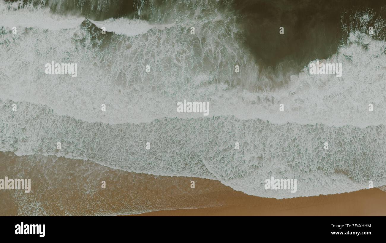 Vista aerea che guarda verso le onde dell'oceano che si infrangono su una spiaggia sabbiosa, con motivi di schiuma marina e texture di acqua vorticose nei toni del verde e del marrone Foto Stock