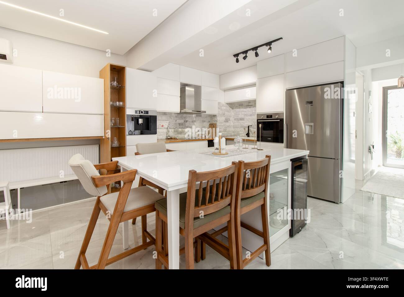 Cucina e zona pranzo minimalista e moderna bianca con sgabelli da bar in legno, elettrodomestici in acciaio inossidabile, isola della cucina e arredamento contemporaneo Foto Stock