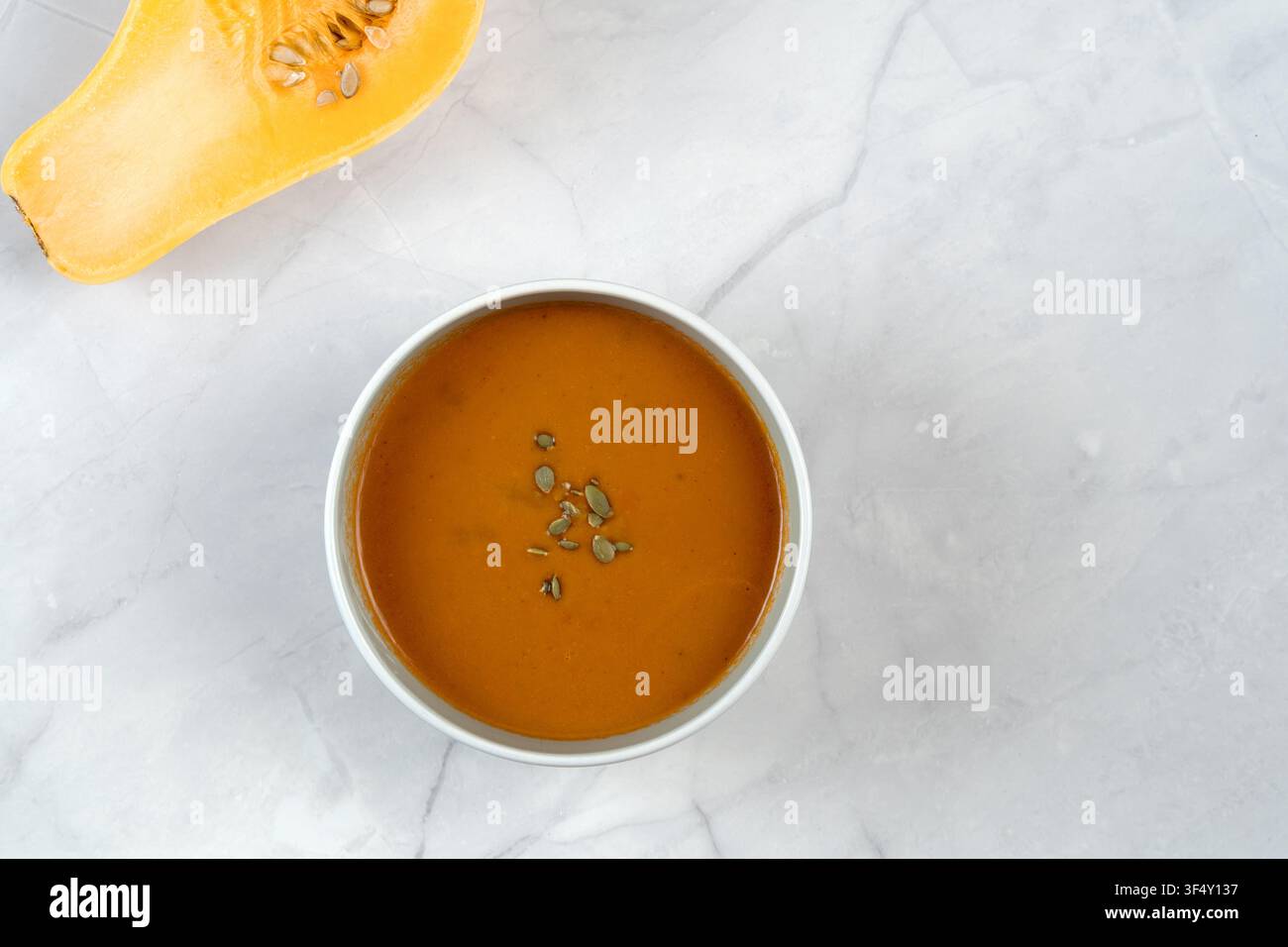 Flat lay di vellutata di zucca arancione liscia cosparsa di semi verdi in ciotola bianca su superficie di marmo chiaro. Stile alimentare minimalista moderno Foto Stock