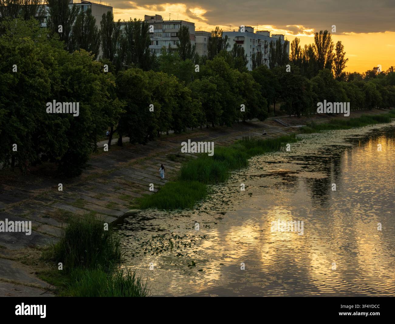 Il fiume Dnepr che scorre lungo un argine di cemento fiancheggiato da alberi e edifici residenziali sotto un cielo illuminato da un tramonto dorato, riflettendo la luce su Foto Stock