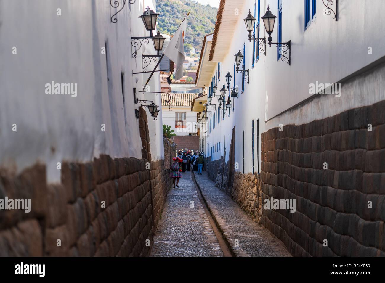 Vista di una stretta strada storica con muri in muratura di pietra tradizionali Inca, edifici coloniali bianchi, lanterne sospese e una persona in abiti tradizionali Foto Stock
