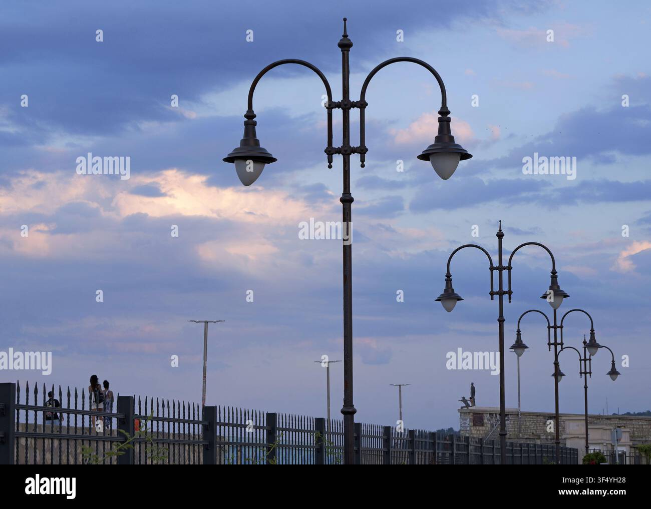 Lampioni ornati delimitano una passeggiata urbana sul lungomare sotto un cielo nuvoloso drammatico, creando un'atmosfera serena serale con luce soffusa Foto Stock
