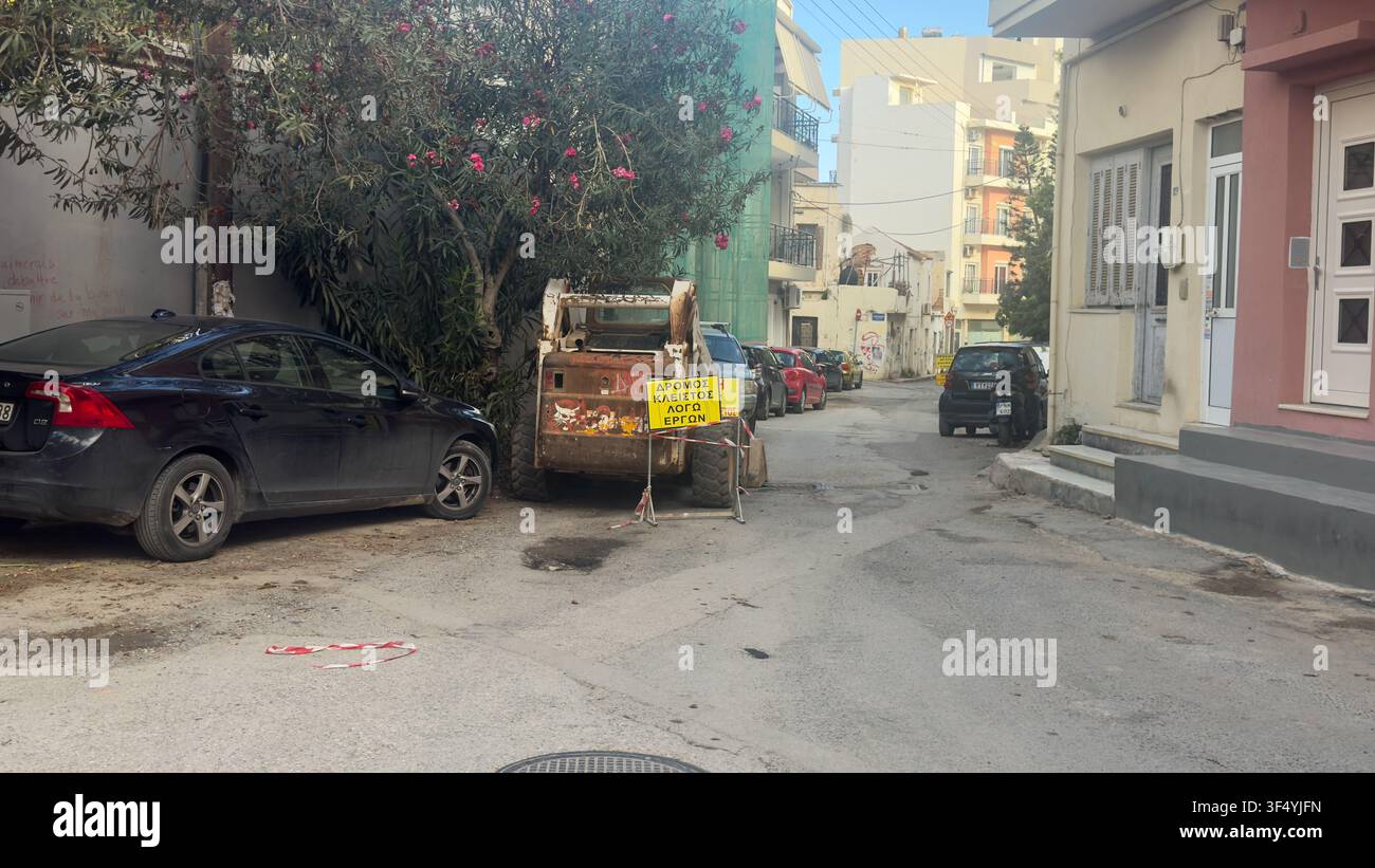 Pesanti macchinari edili bloccano una strada residenziale con auto parcheggiate e un cartello greco di cantiere che dice "Strada chiusa per lavori" Foto Stock