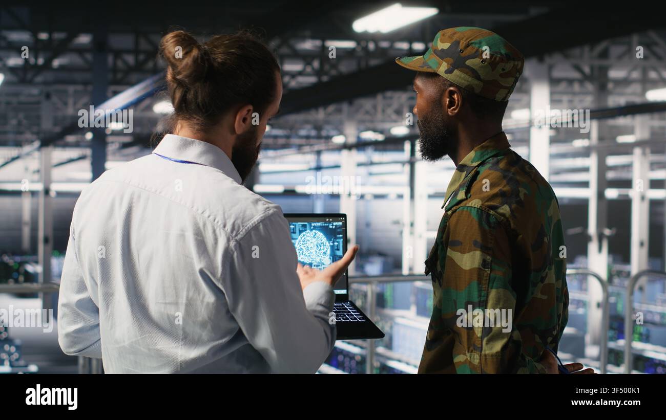 I tecnici militari in un data center sicuro utilizzano un notebook per monitorare i sistemi di intelligenza artificiale. Operatori dell'esercito in un hub server che garantisce un'infrastruttura stabile per le operazioni di apprendimento automatico Foto Stock