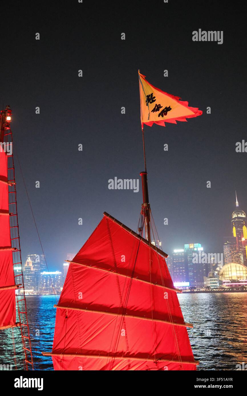 Prima verticale di una vela tradizionale di giunco rosso e di uno stendardo sventolante di notte con lo skyline illuminato di Victoria Harbour, Hong Kong. Foto Stock