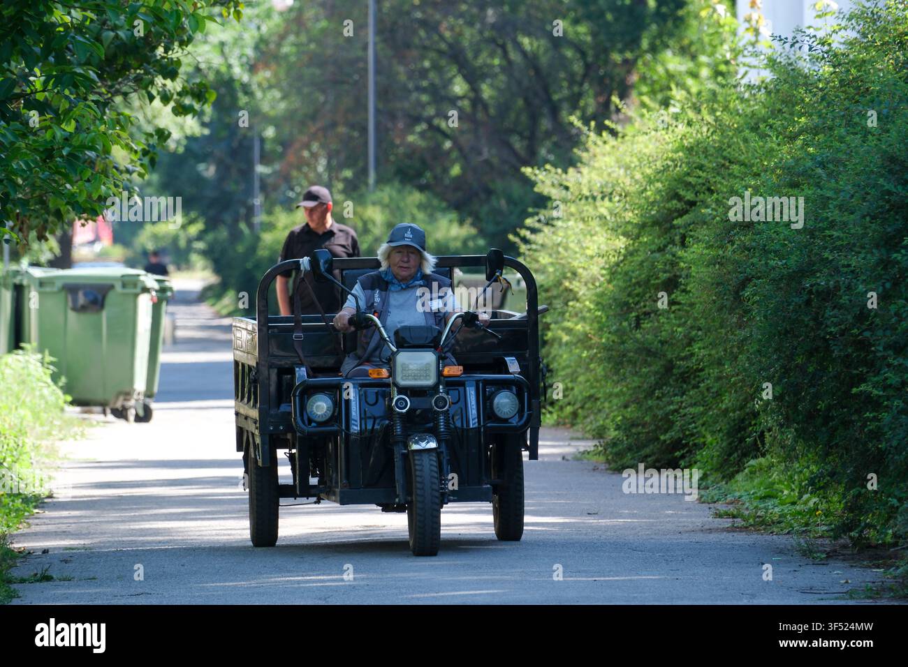 KIEV, UCRAINA - 30 luglio 2026: Un veicolo elettrico merci utilitario guidato da un addetto alla manutenzione del parco percorre un sentiero lastricato in un parco cittadino. La scena Foto Stock