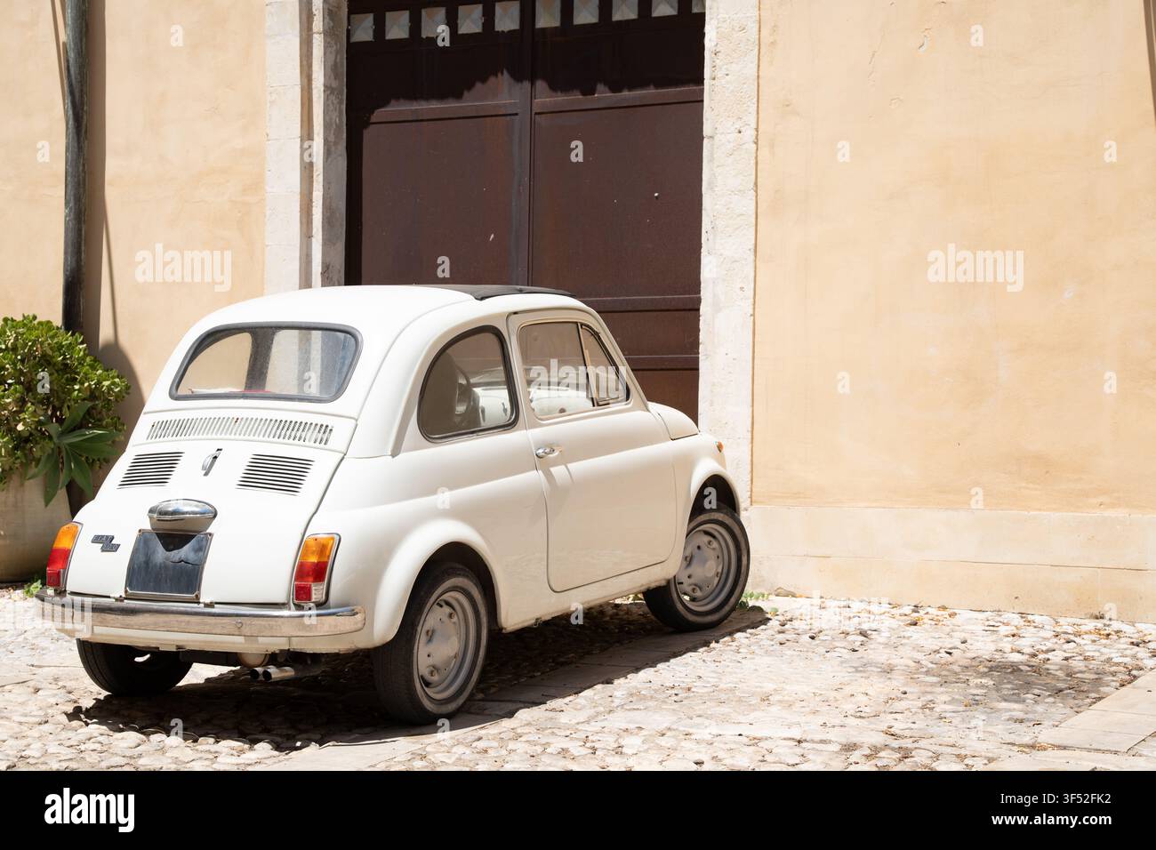 Fiat 500 bianco vintage parcheggiato in un cortile lastricato di edifici in pietra giallo chiaro in Sicilia Foto Stock