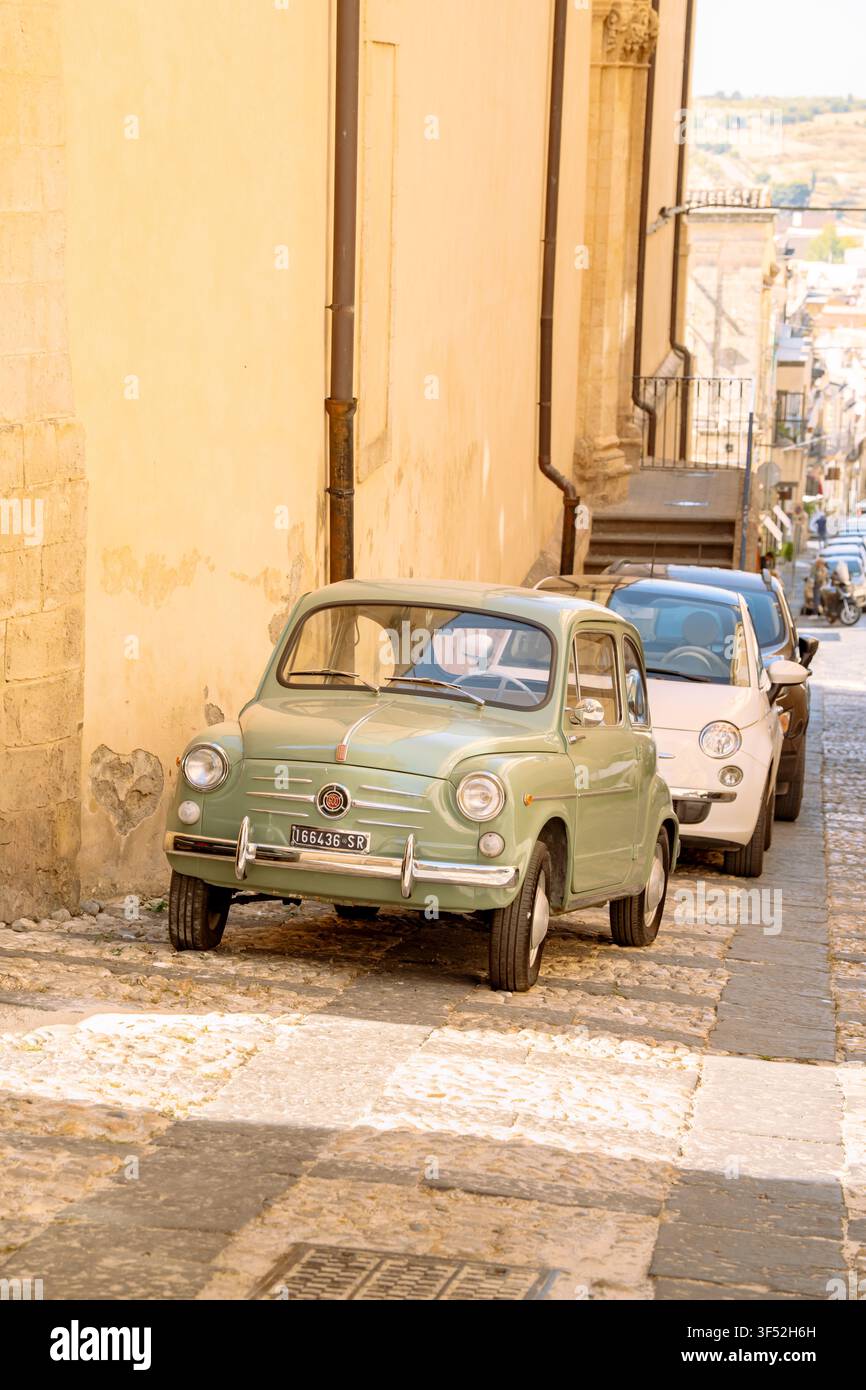Fiat 600 vintage verde, una versione più grande della 500, parcheggiata su una strada acciottolata di edifici in pietra giallo chiaro in Sicilia Foto Stock